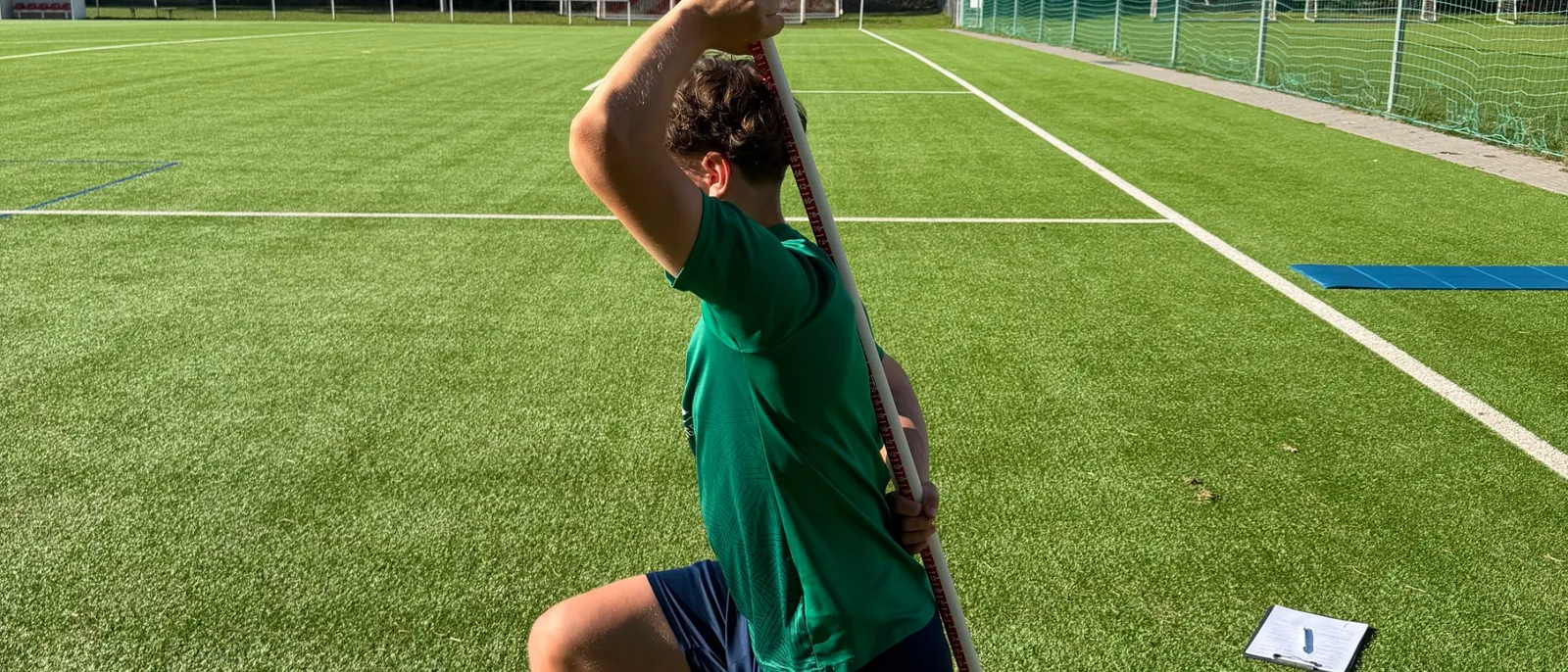 Spieler im grünen Trikot von schräg hinten im Kniestand, er hält einen langen Stab senkrecht hinter dem Rücken, im Hintergrund ein sonniger Kunstrasenplatz