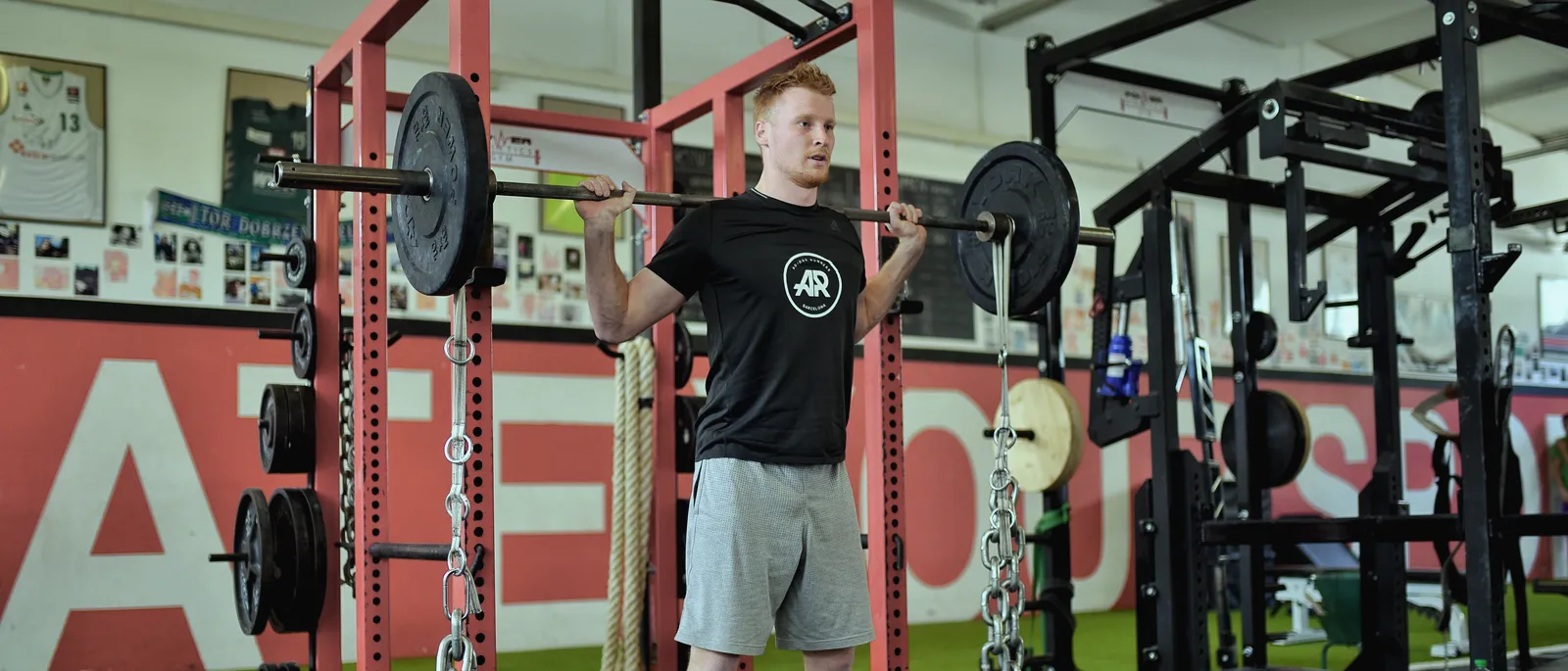 Athlet steht mit der Langhantel auf den Schultern in einem roten Power Rack im Kraftraum, an den Enden der Stange hängen Ketten
