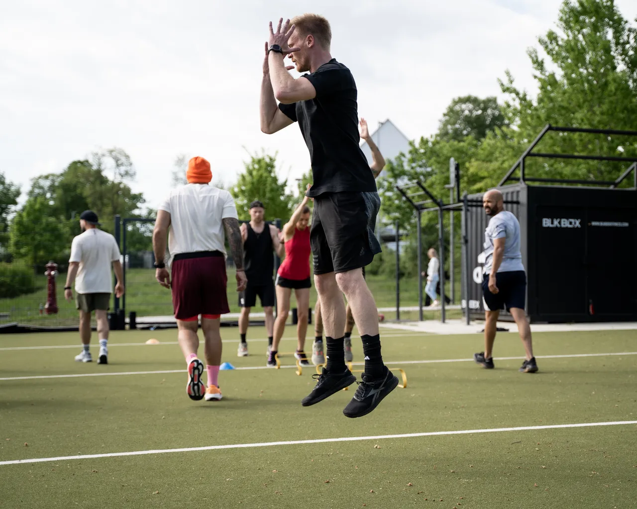 Daniel Hense in der Luft bei einem explosiven Sprung auf dem Kunstrasen, beide Füße vom Boden, im Hintergrund eine Trainingsgruppe beim Aufwärmen mit gelben Minihürden