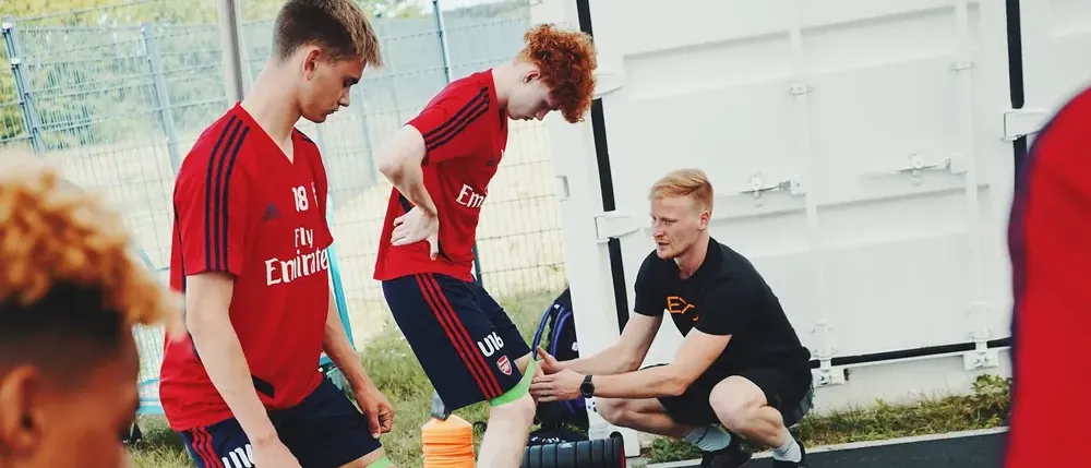 Daniel Hense hockt neben einem Nachwuchsspieler im roten Trikot und korrigiert mit den Händen dessen Kniestellung bei einer Übung mit Miniband, links daneben macht ein zweiter Spieler dieselbe Übung